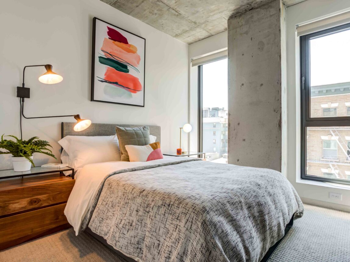 Luxury Apartments in Downtown San Francisco, CA - 50 Jones - Bedroom with Carpet Flooring, Bed, Nightstands with Lamps, Exposed Concrete Ceiling, and Floor-to-Ceiling Windows.