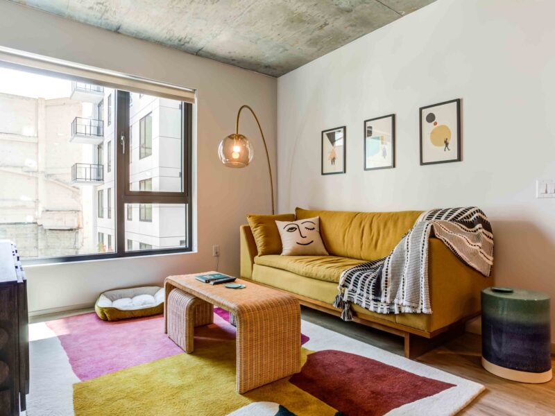 High-End Apartments in Downtown San Francisco, CA - 50 Jones - Living Room with Luxury Wood-Style Vinyl Plank Flooring, Area Rug, Yellow Sofa, Coffee Table with Pull-Out Stool, Floor Lamp, Exposed Concrete Ceiling, and Large Window.