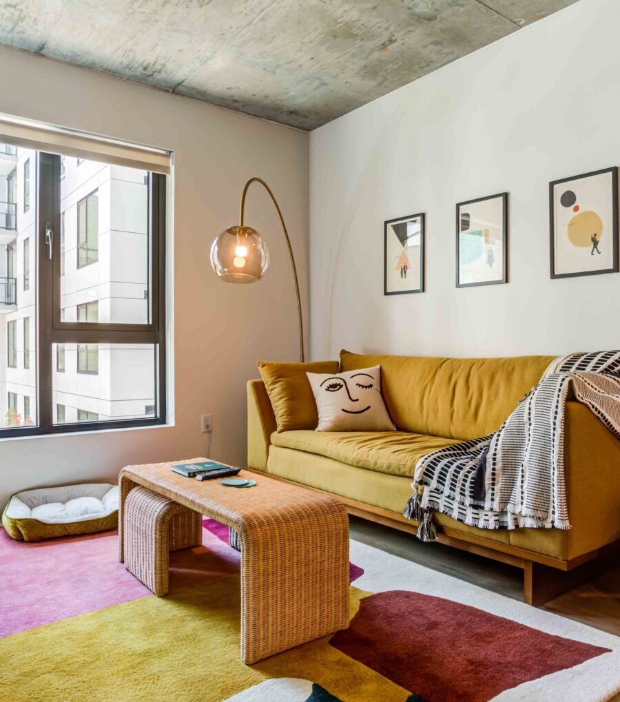 High-End Apartments in Downtown San Francisco, CA - 50 Jones - Living Room with Luxury Wood-Style Vinyl Plank Flooring, Area Rug, Yellow Sofa, Coffee Table with Pull-Out Stool, Floor Lamp, Exposed Concrete Ceiling, and Large Window.